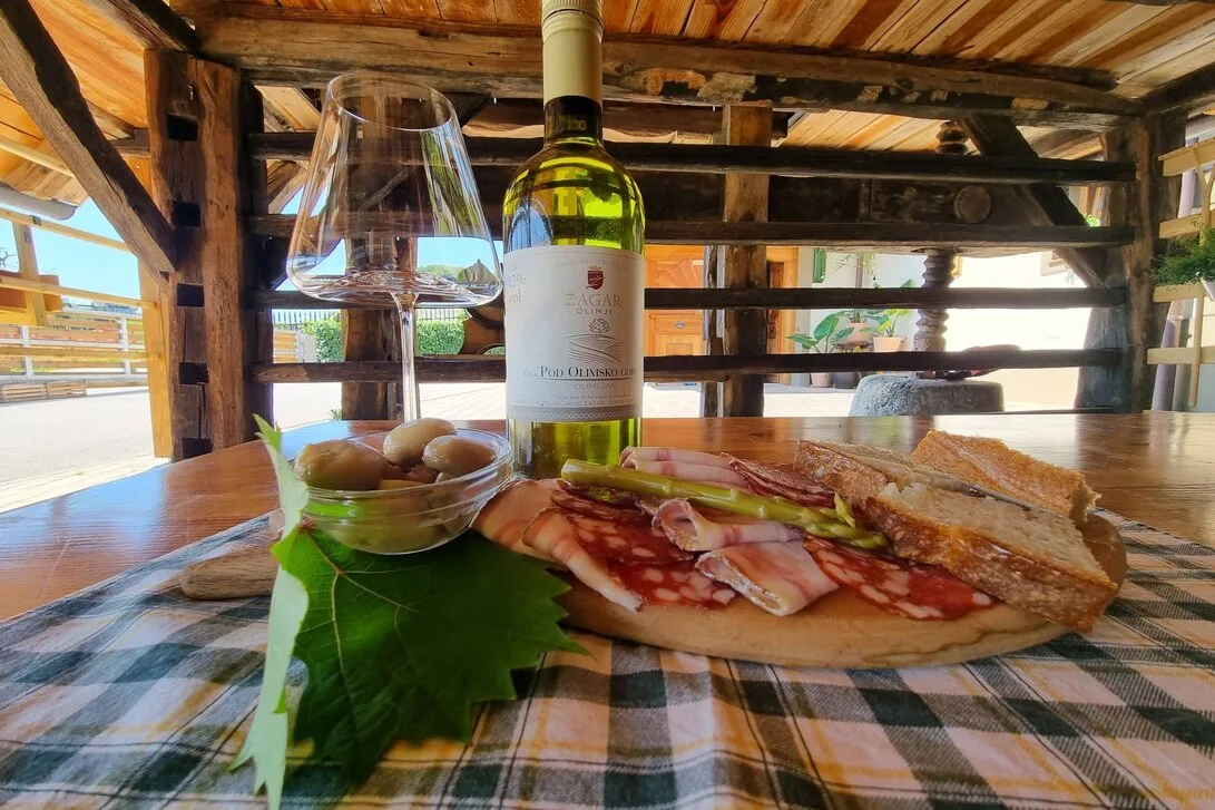 Steklenica belega vina Žagar s kozarcem, narezkom, kruhom in olivami na karirastem prtu pod prešo.